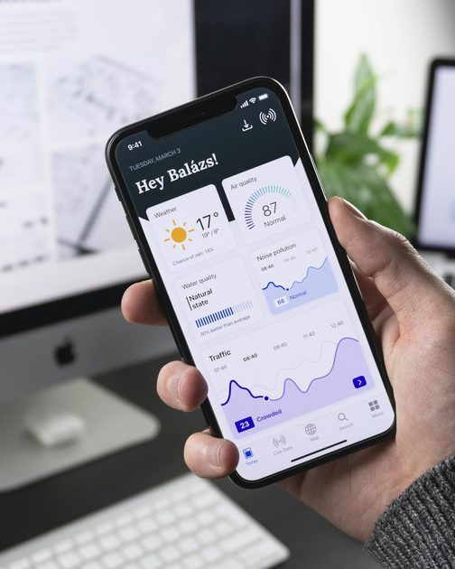 A hand holding a phone showing a dashboard of charts in front of a desktop screen