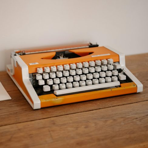 An orange typewriter beside a blank sheet of paper on a desk