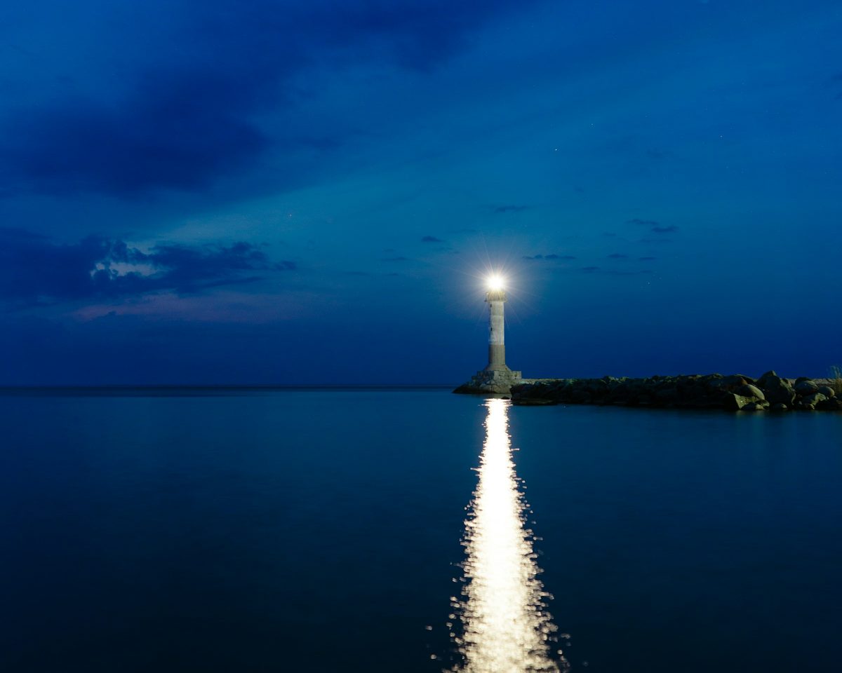 A single light at the end of a pier, reflected in dark blue water at dusk