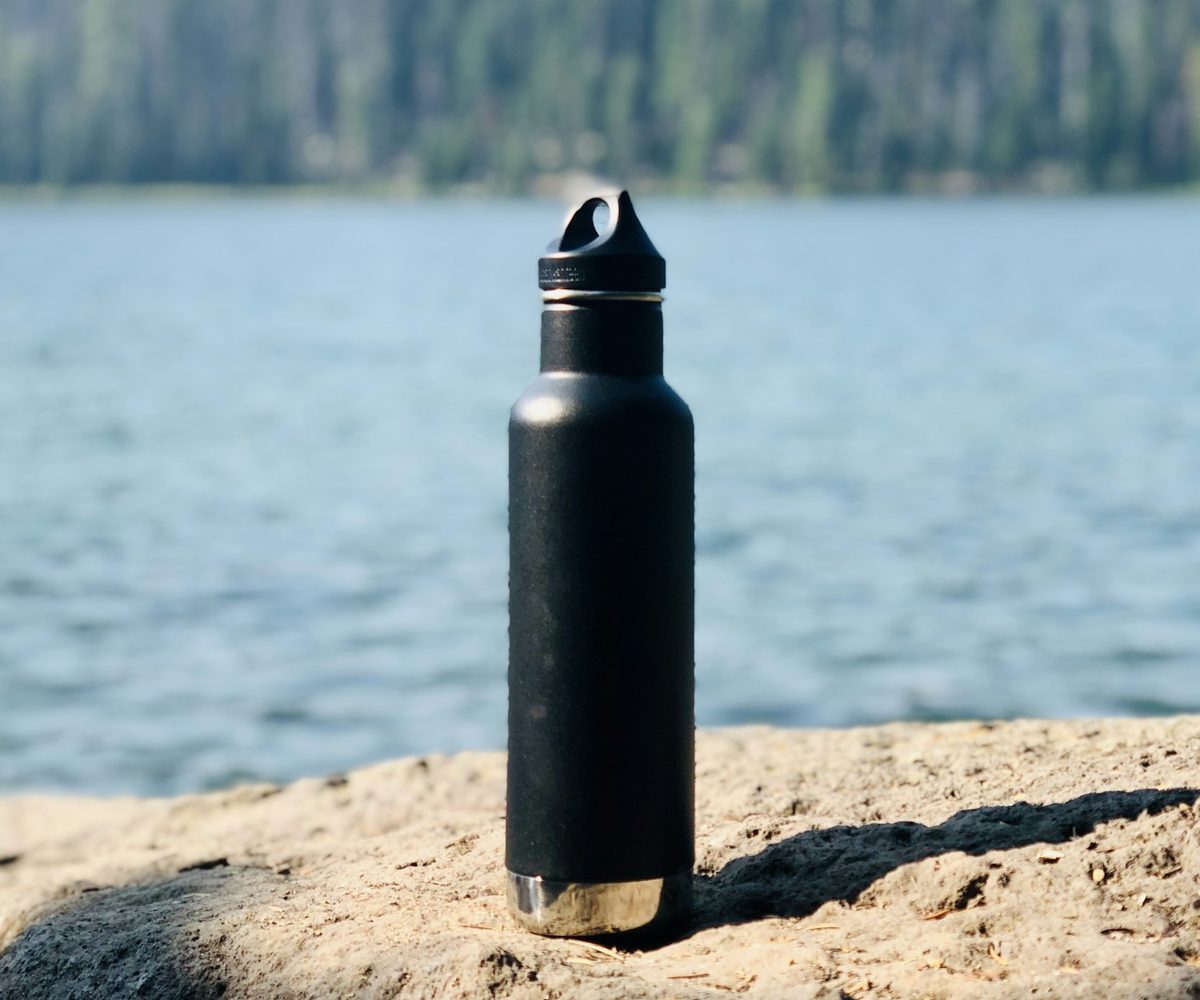 A matte dark water bottle standing on a rock by a lake