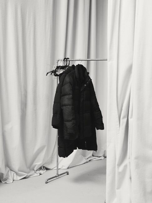 A black puffer coat on a clothing rack against pale curtains