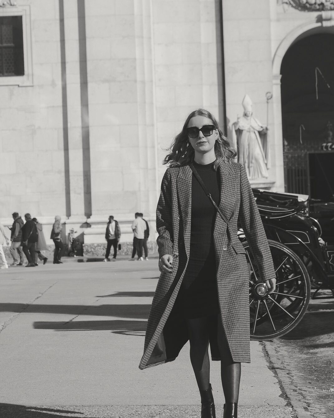 A woman in a long coat and sunglasses walking past a stone building