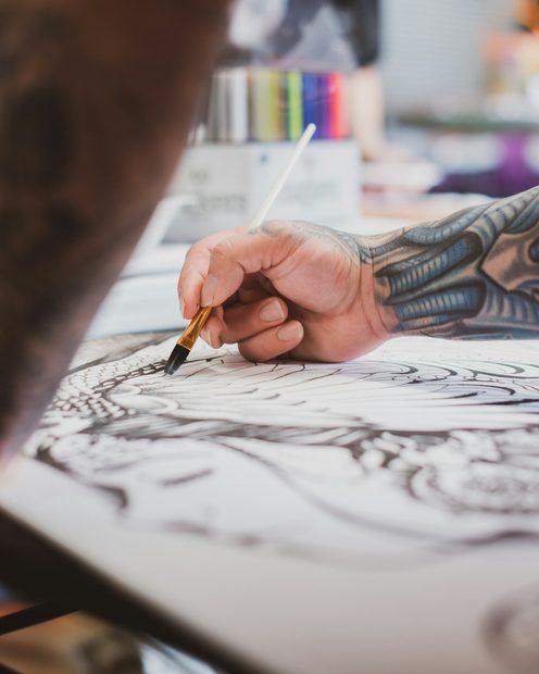 A tattooed hand inking a detailed design on paper