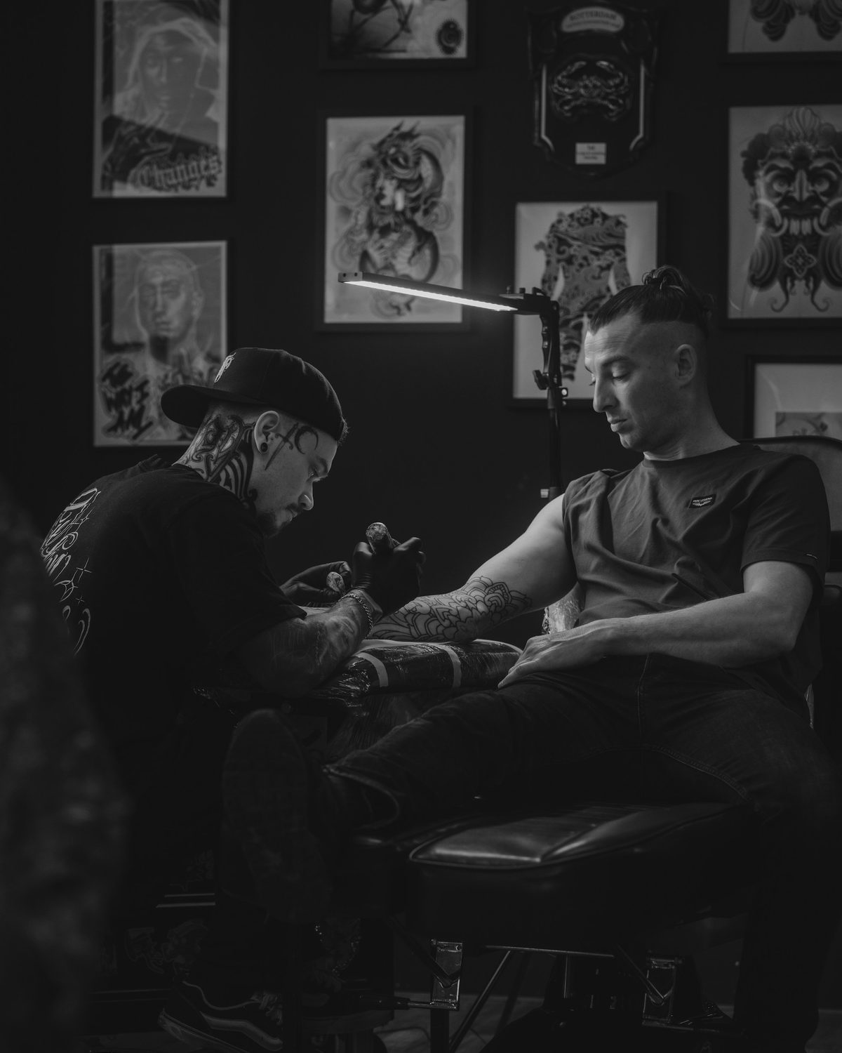 A tattoo artist working on a client's forearm in a dark studio hung with framed flash