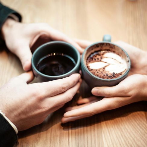 Two people holding cups of coffee across a wooden table