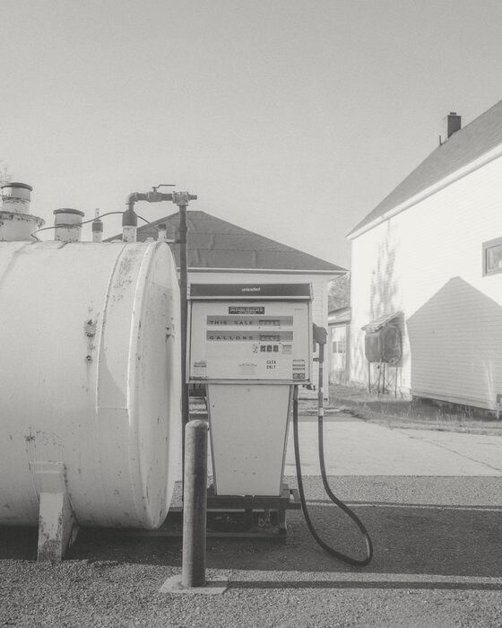 A lone fuel pump beside a white tank and a white house