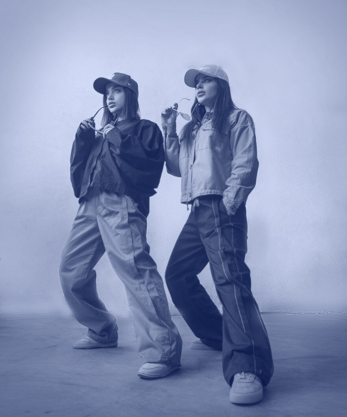 Two models in a studio, one in a black bomber and one in a green overshirt, both in caps