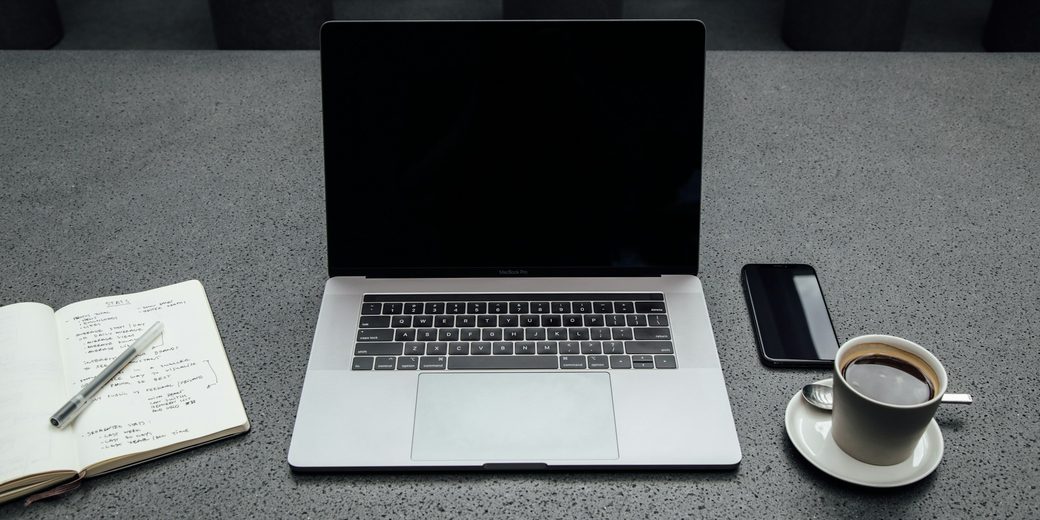 A laptop, open notebook, phone and coffee on a gray desk, seen from above
