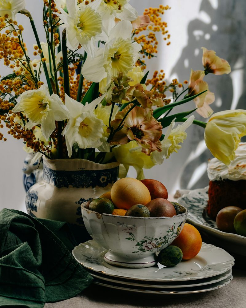 Daffodils in a blue and white jug beside a bowl of eggs and fruit