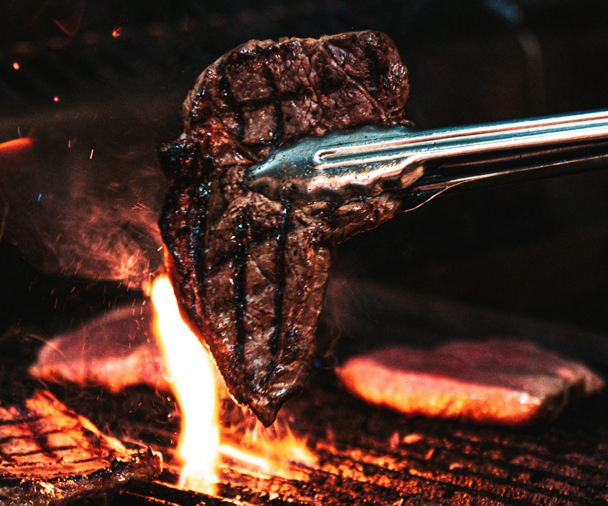 A steak lifted with tongs over a flaming charcoal grill