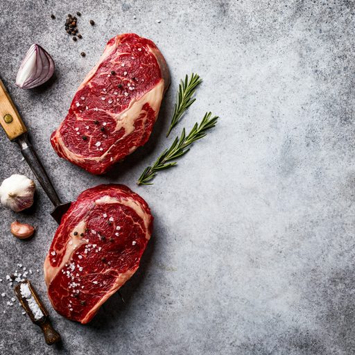 Two thick ribeye steaks on a gray stone with rosemary and garlic