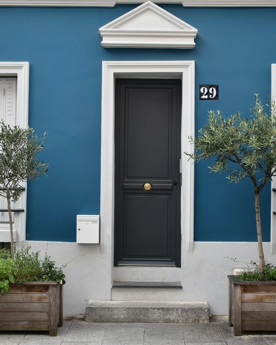 A dark front door set in a blue house with olive trees in planters