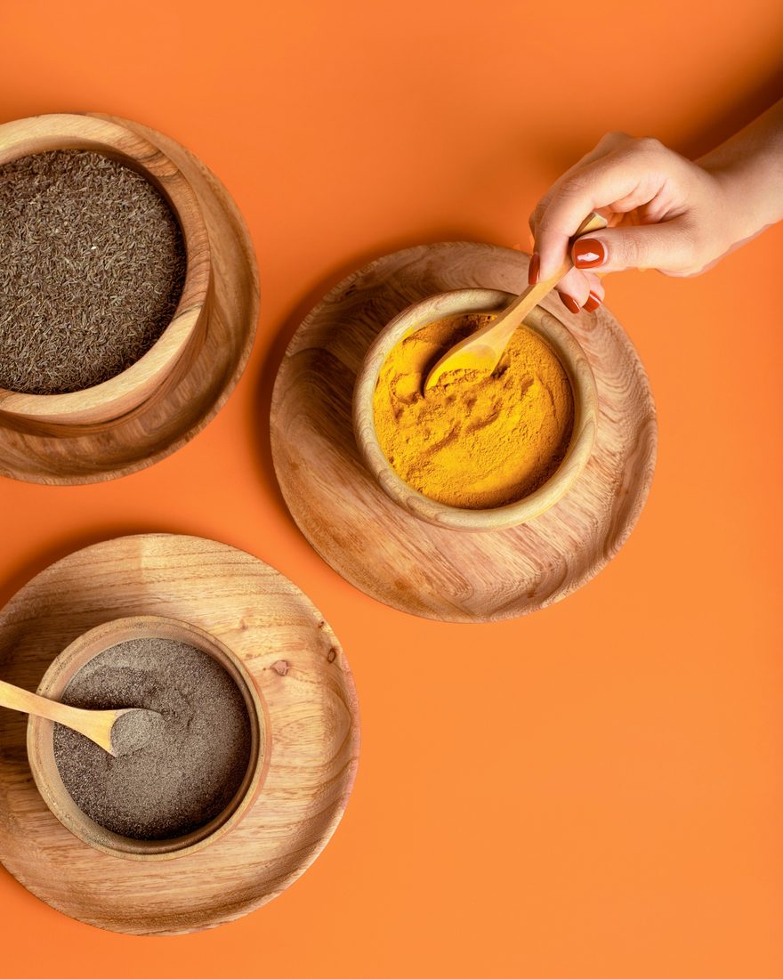 A hand stirring turmeric in a wooden bowl beside bowls of cumin and pepper