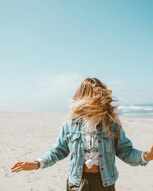 A woman in a denim jacket walking barefoot along a pale beach