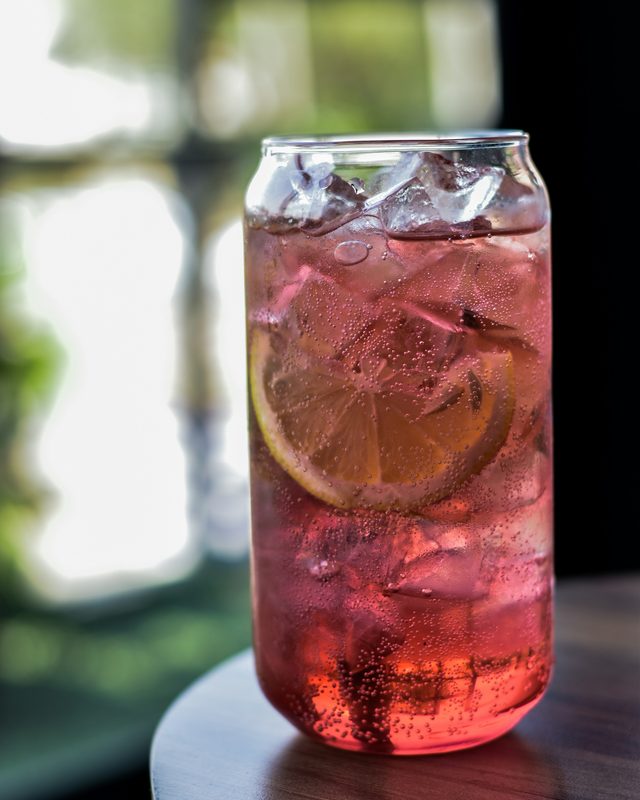 A tall glass of dark berry soda over ice with a lemon wheel
