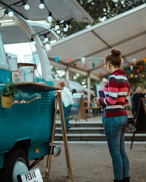 A woman in a striped sweater waiting at a teal food truck under string lights