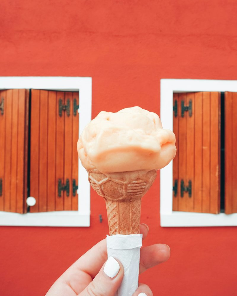 A hand holding a peach gelato cone against a red wall with wooden shutters