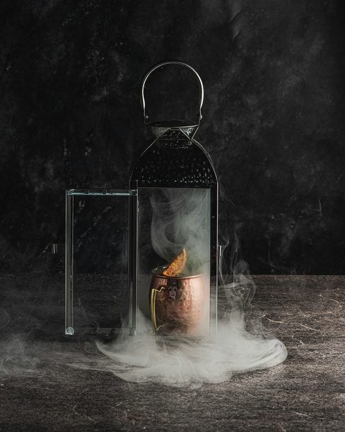 A smoking cocktail inside a glass lantern on a dark stone bar