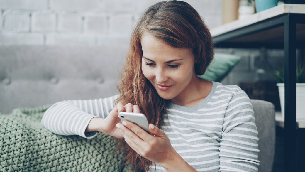 A smiling woman on a sofa replying to a message on her phone