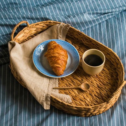 A croissant and a cup of coffee on a wicker tray on a striped bed