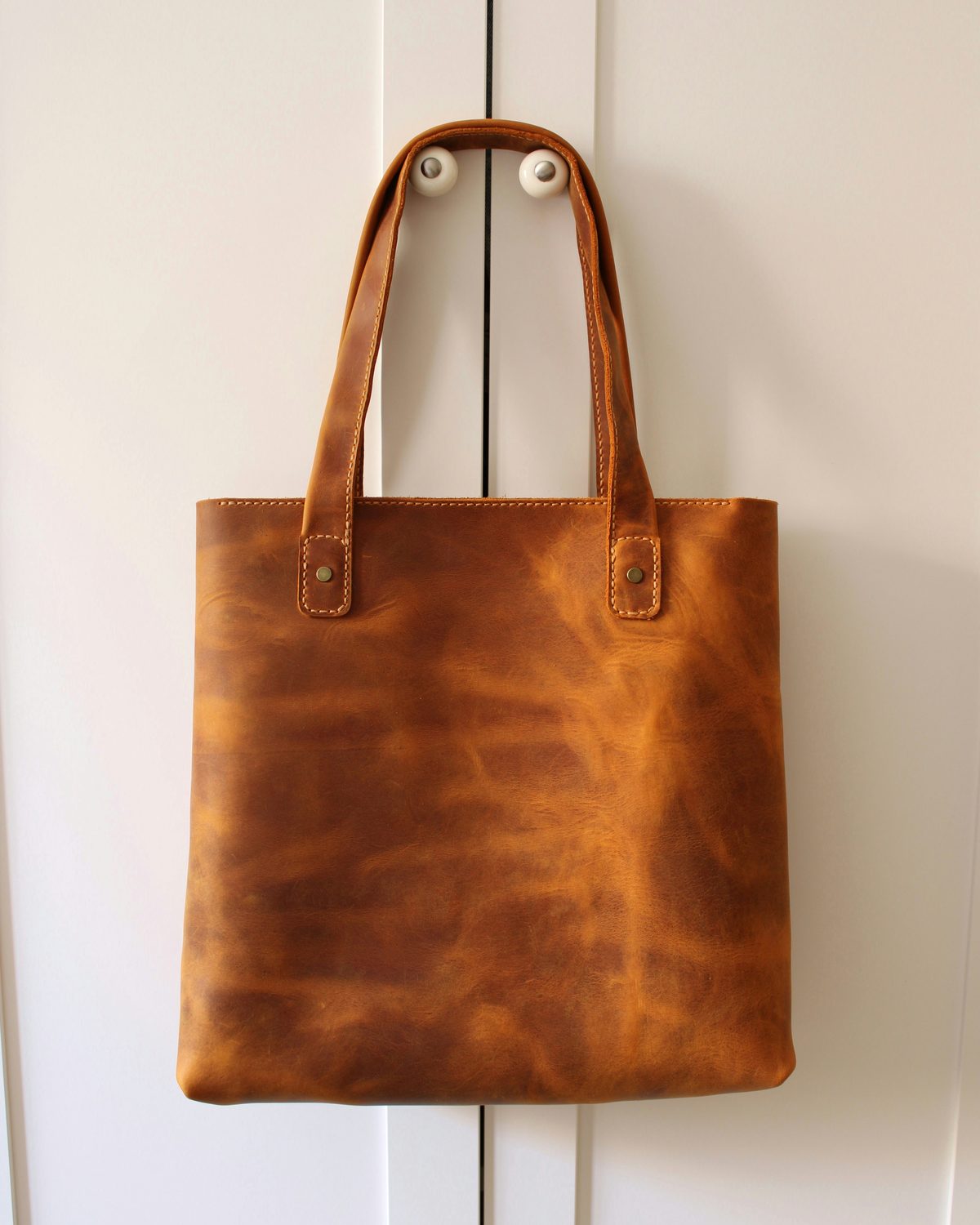 A tan leather tote hanging from a peg against a white wall
