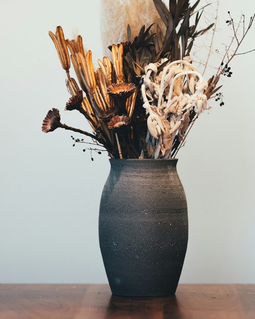 A dark stoneware vase of dried flowers on a wooden table