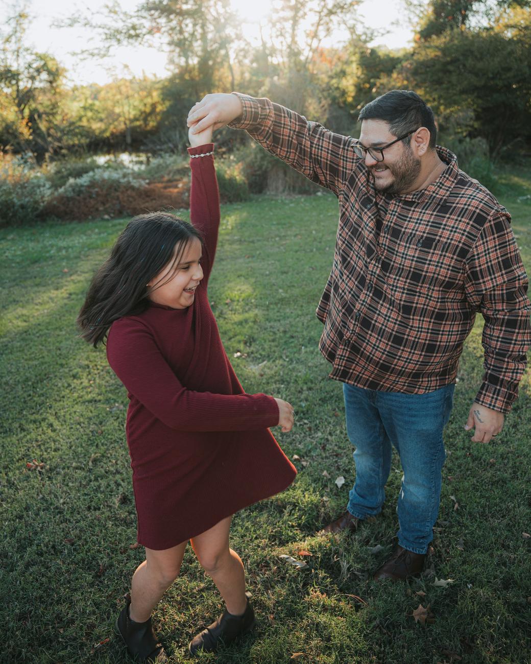 A father twirling his daughter in a park, low sun through the trees