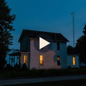 A white farmhouse with its lights on at dusk, with a play button