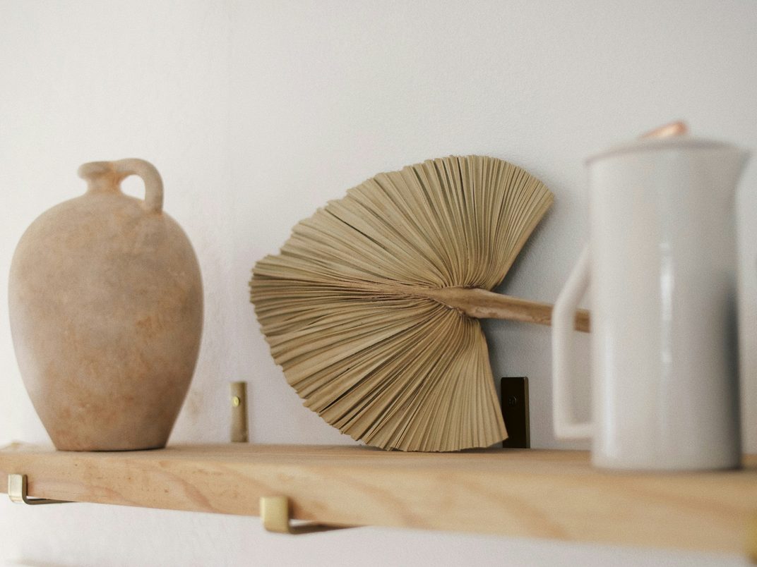 A clay vase, a woven palm fan and a white jug on a pale wooden shelf