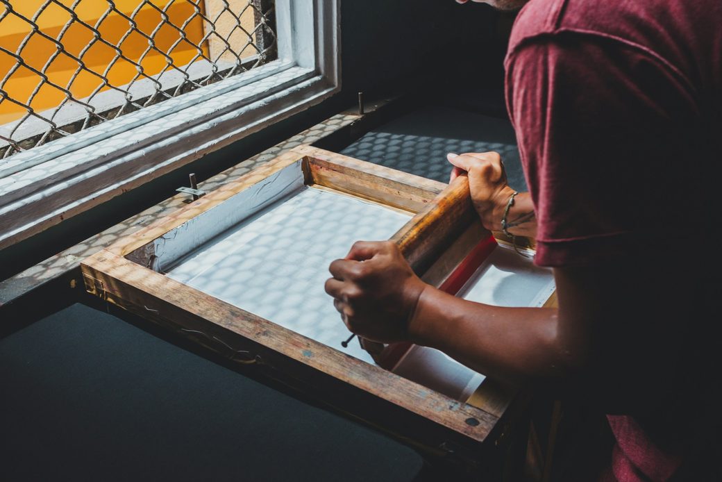 An artist pulling a fresh print through a silkscreen frame by a workshop window