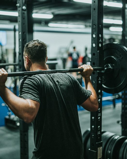 A man in a black t-shirt with a barbell across his back in a squat rack