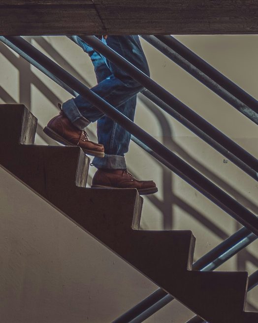 Legs in jeans and work boots climbing a concrete stairwell beside a brick wall