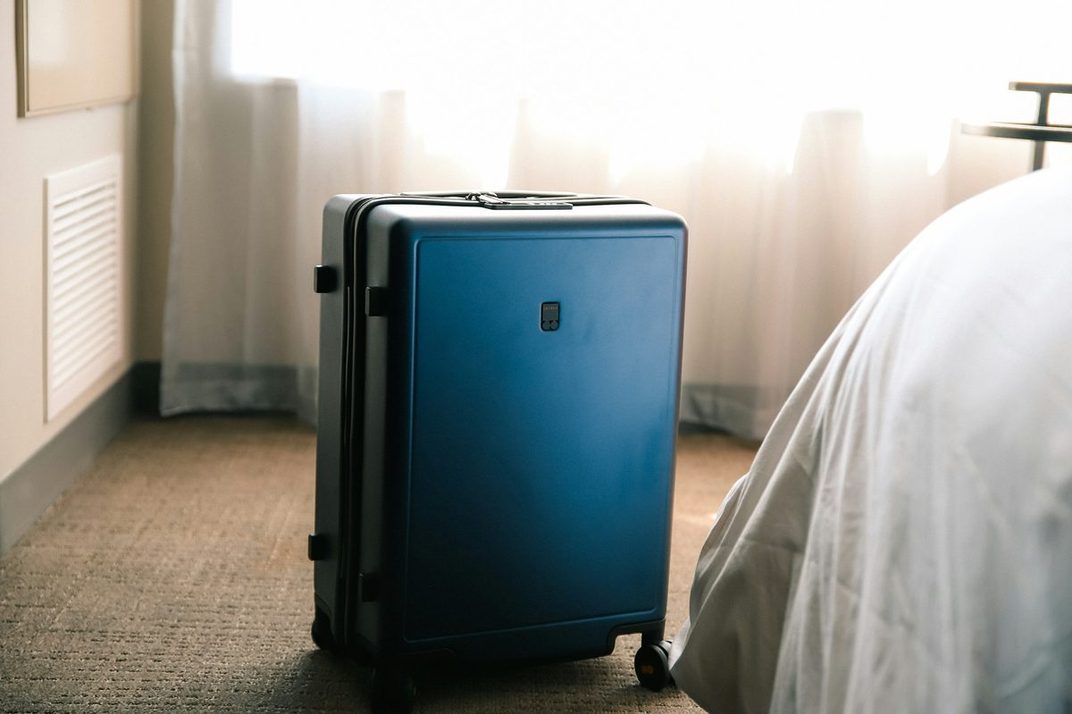 A blue hardshell suitcase standing beside a bed in a sunlit room