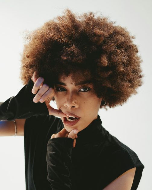 A woman with a full, rounded afro against a pale wall