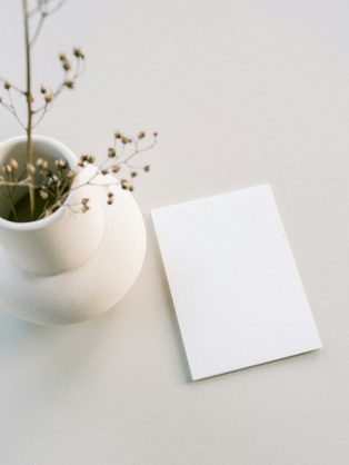 A blank card leaning beside a small white vase of dried stems