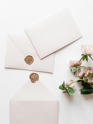 Blush envelopes closed with gold wax seals beside pink flowers