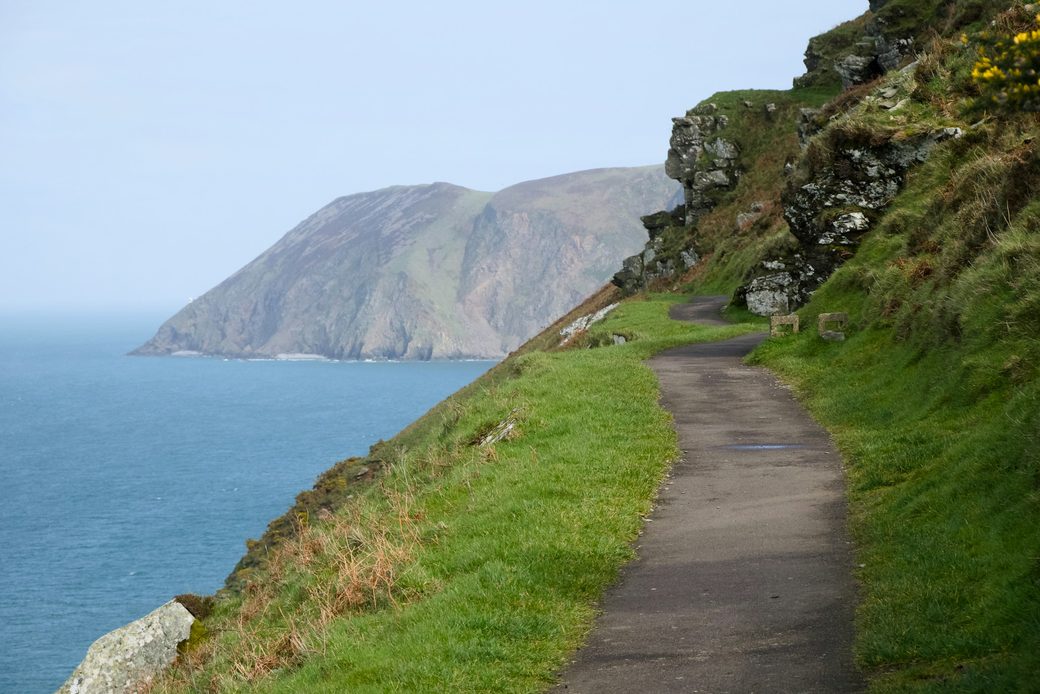 A narrow path along a grassy cliff, with a far headland across the water