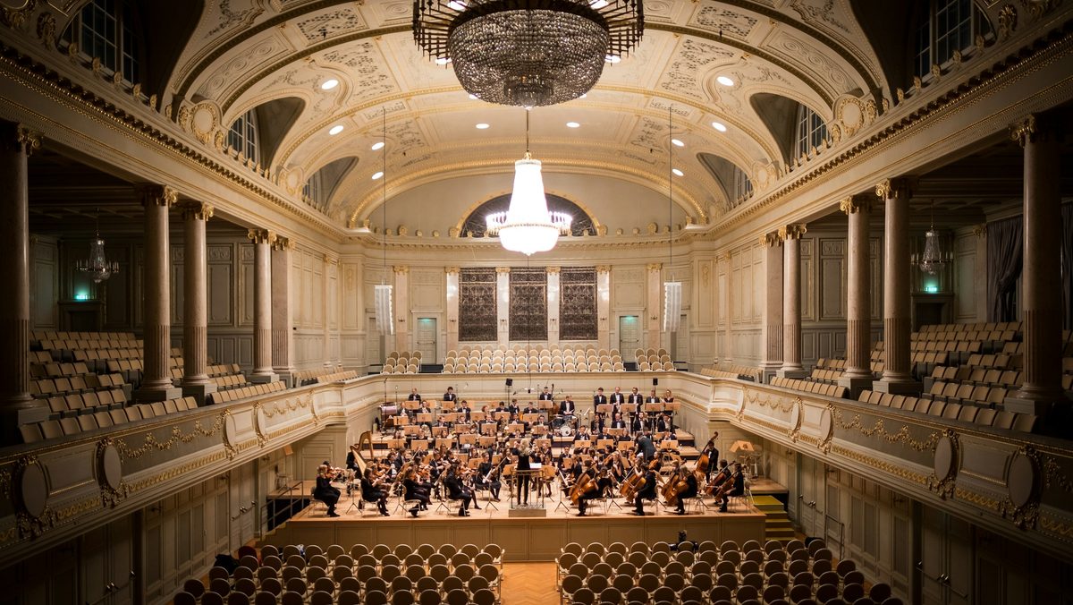 An orchestra on stage in a grand concert hall under chandeliers