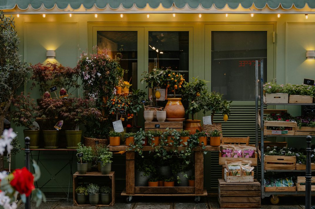 A small flower shop under a green awning, its plants spilling onto the sidewalk under warm lights