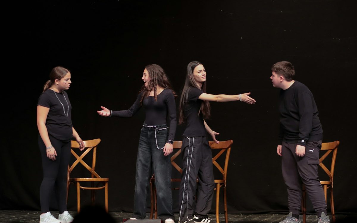 Four actors rehearsing a scene on a dark stage with chairs