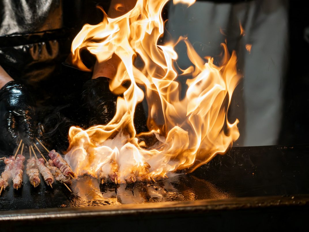 Skewers of meat over a roaring grill flame at a food stall
