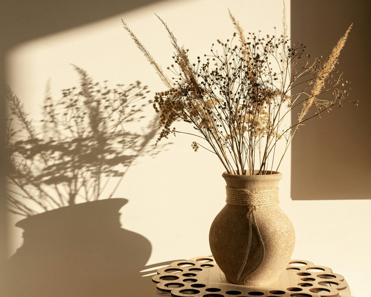 A clay vase of dried grasses on a lace mat, casting a long shadow on a warm wall