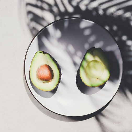Two avocado halves on a white plate under a palm leaf shadow