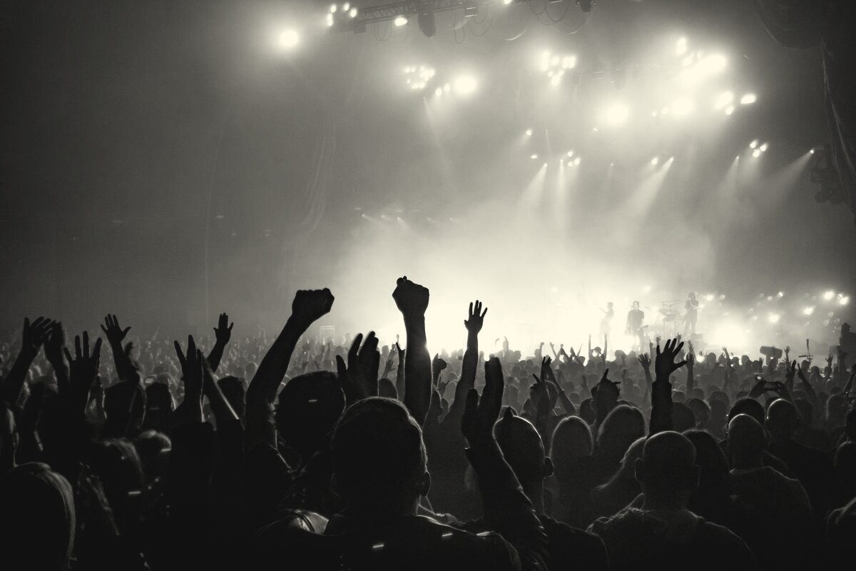 A crowd with raised hands facing a lit stage, in black and white