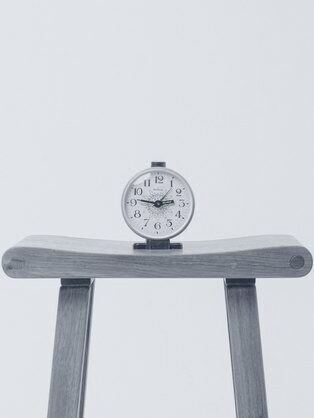 A small brass clock on a wooden stool against a pale wall