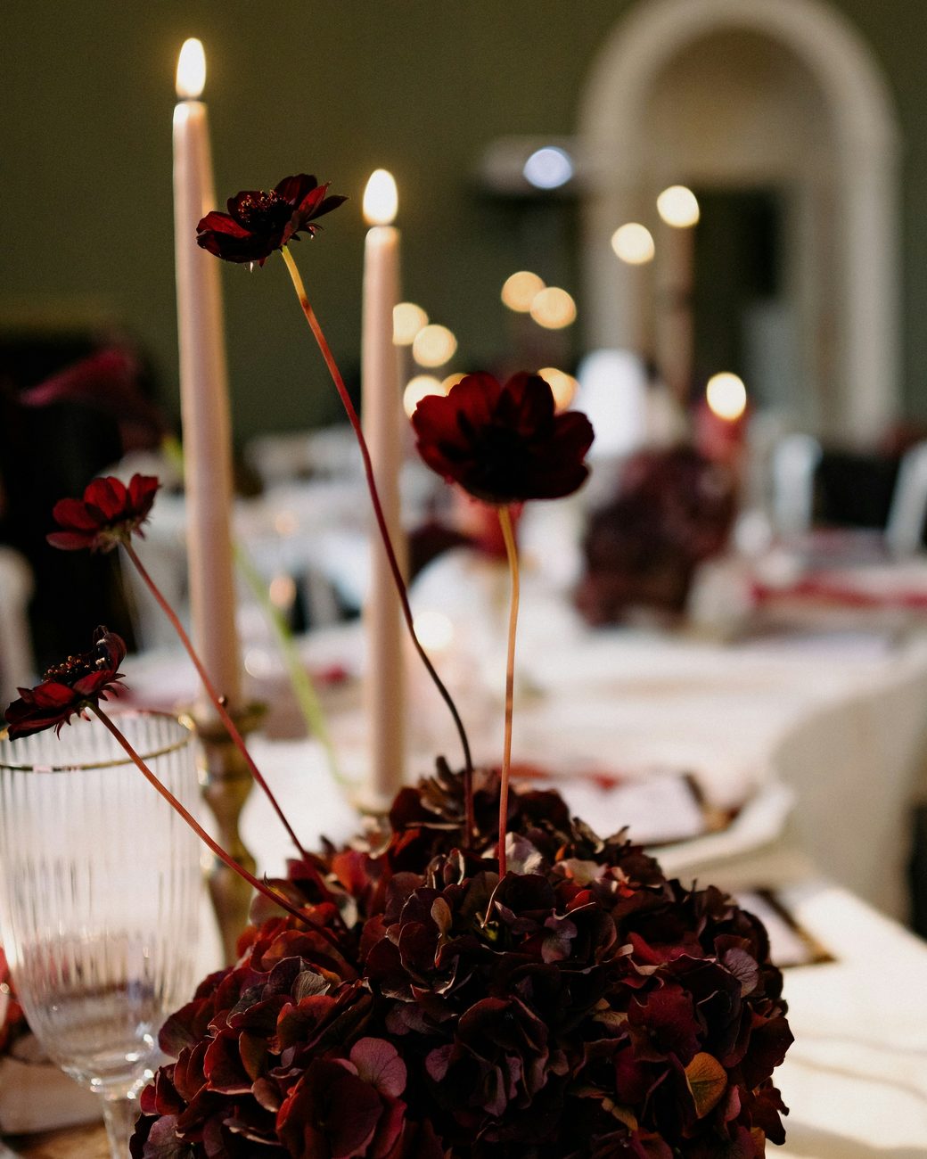 Tall lit tapers and dark red flowers on a set table in a dim dining room