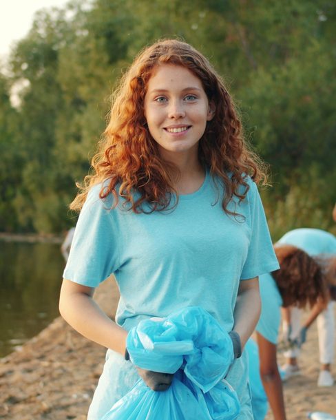 A smiling volunteer holding a full bag by a lake, with others picking up litter behind her