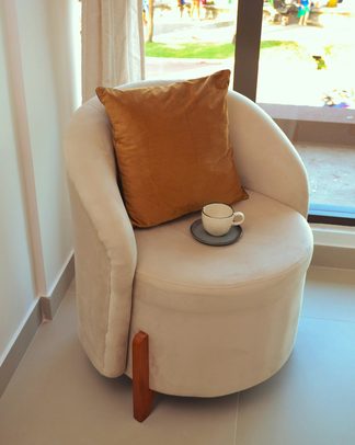 A cream curved armchair with a rust cushion by a window