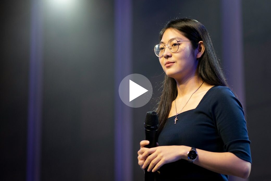 Maya Castell speaking on a dark stage, with a play button over the frame