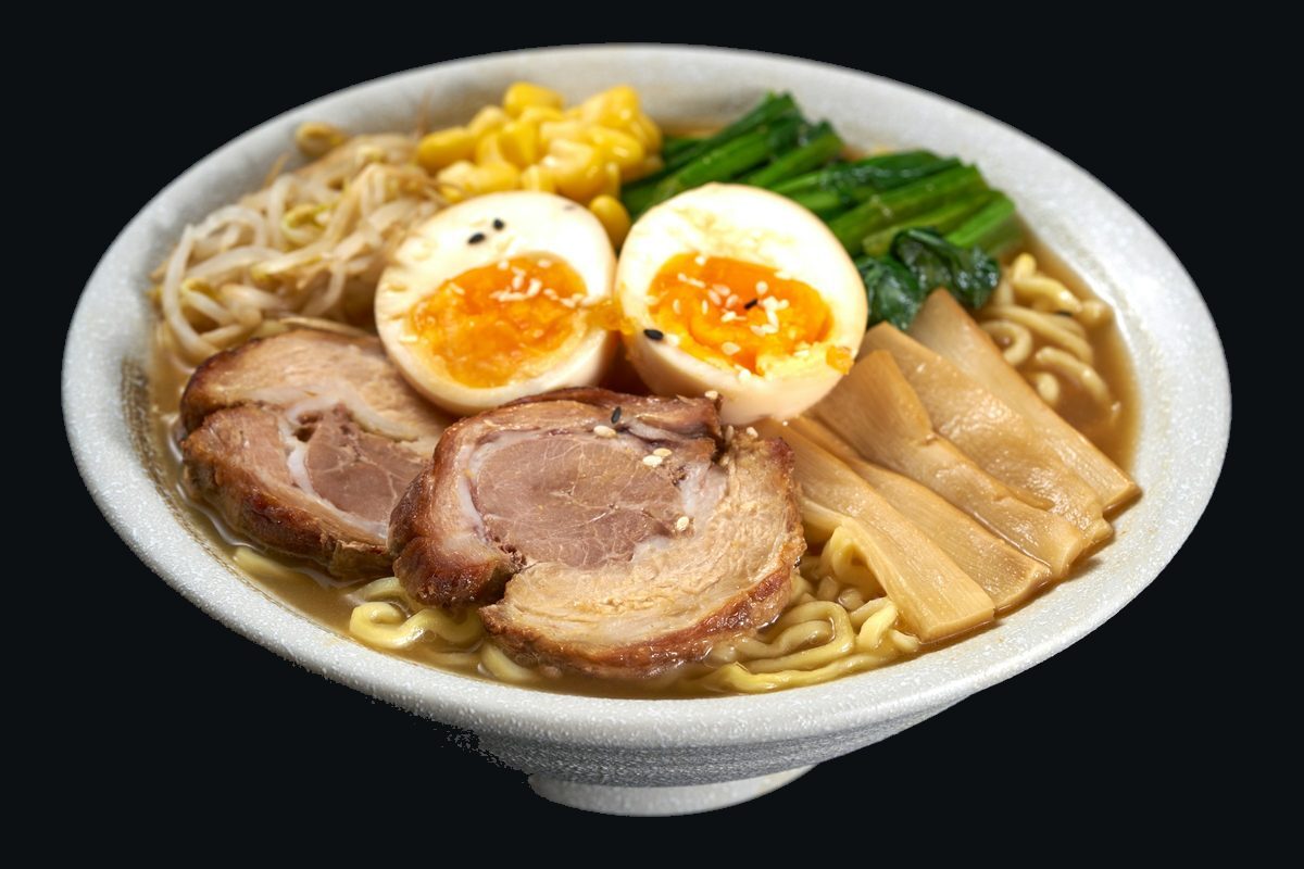 A bowl of ramen with pork, soft eggs, greens and noodles on a dark table